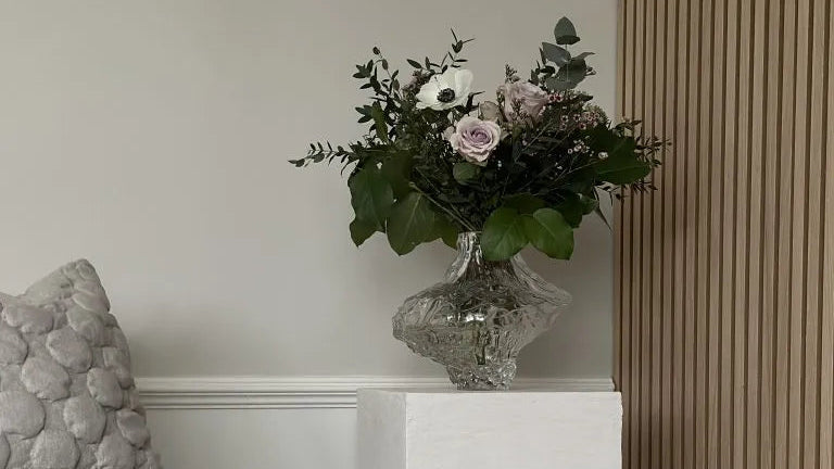 Glass vase with bouquet on white pedestal, next to a textured gray pillow and Akupanel acoustic wood panel wall for a natural Nordic setting.